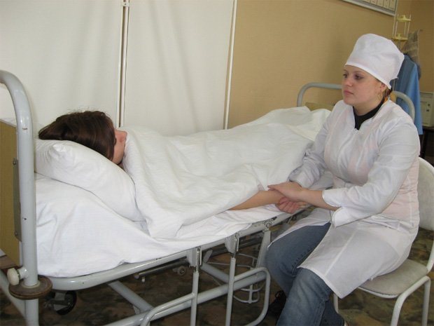 У кровати больной девушки сидит медицинская сестра и держит ее за руку