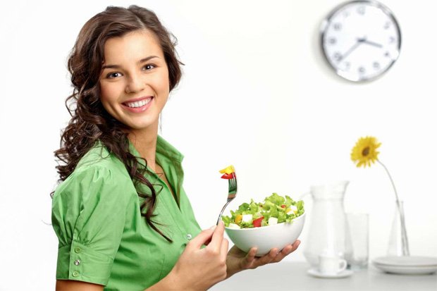 Улыбающаяся девушка на фоне часов ест полезный салат из миски