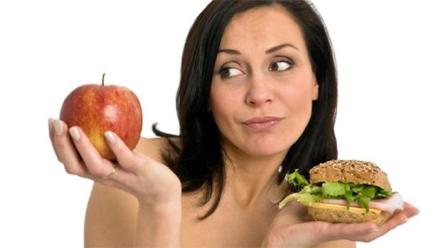 Девушка держит в одной руке бургер, а в другой яблоко и смотрит на него