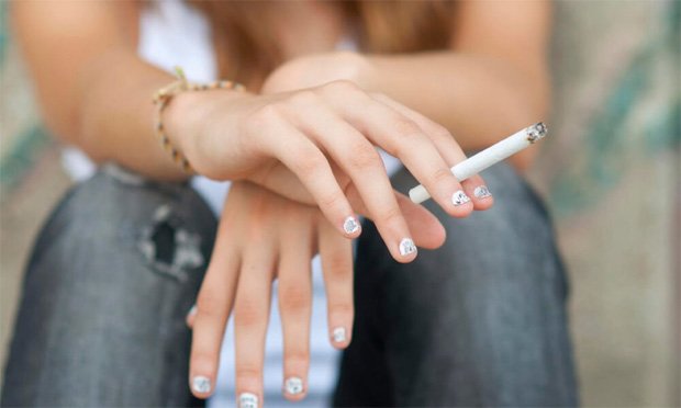 Девушка в джинсах сидит и держит зажженную сигарету в руках
